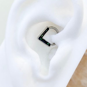 May include: A silver hoop earring with a black and green square-shaped charm. The charm has a glittery finish.