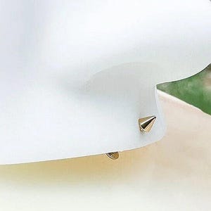 Op de afbeelding: Een close-up van een gouden oorbel met een puntige punt. De oorbel ligt op een wit oppervlak.