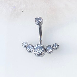 May include: A silver belly button ring with five clear round gems. The gems are arranged in a crescent shape.