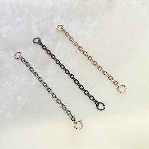 May include: Three metal chain extenders in silver, black, and gold. Each chain has a small ring at both ends, designed for jewellery making or adding length to necklaces and bracelets. The chains are displayed on a light, speckled surface.