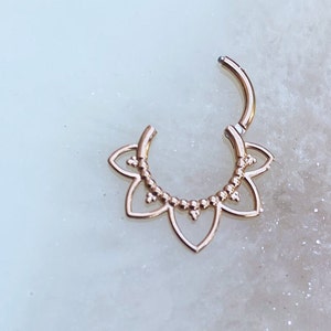 Op de afbeelding: Rosegouden septum clicker ring. De ring heeft een bloemenontwerp met een reeks kleine kralen en bloembladvormige uitsparingen. De clicker is open en toont het sluitmechanisme.