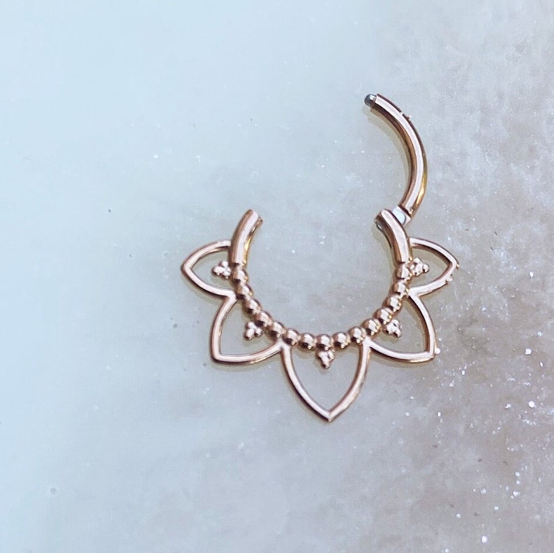 Op de afbeelding: Rosegouden septum clicker ring. De ring heeft een bloemenontwerp met een reeks kleine kralen en bloembladvormige uitsparingen. De clicker is open en toont het sluitmechanisme.