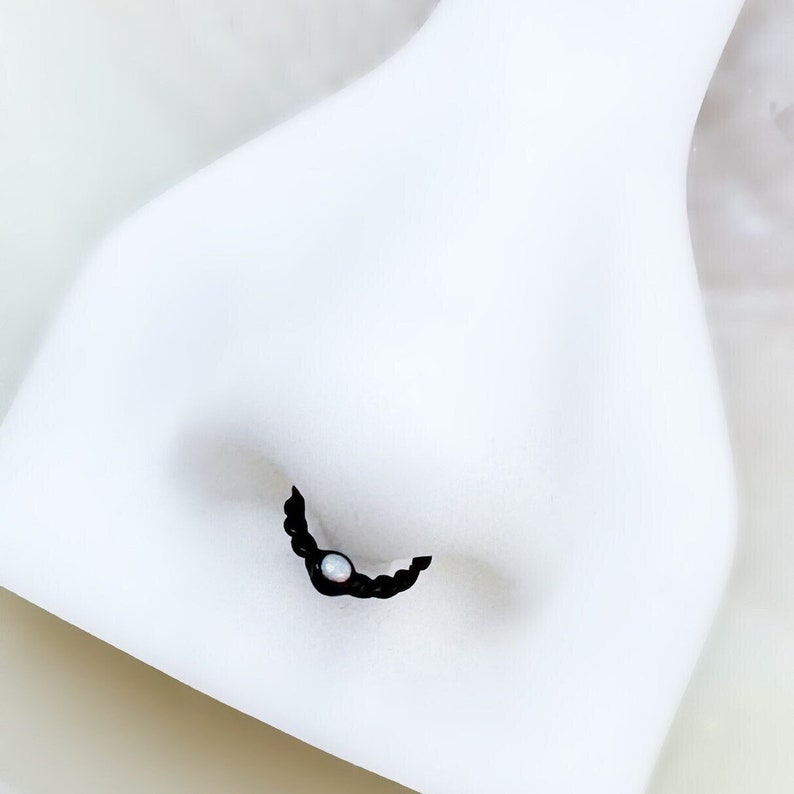 May include: Black nose ring with a small opal gemstone. The ring is shaped like a crescent moon.