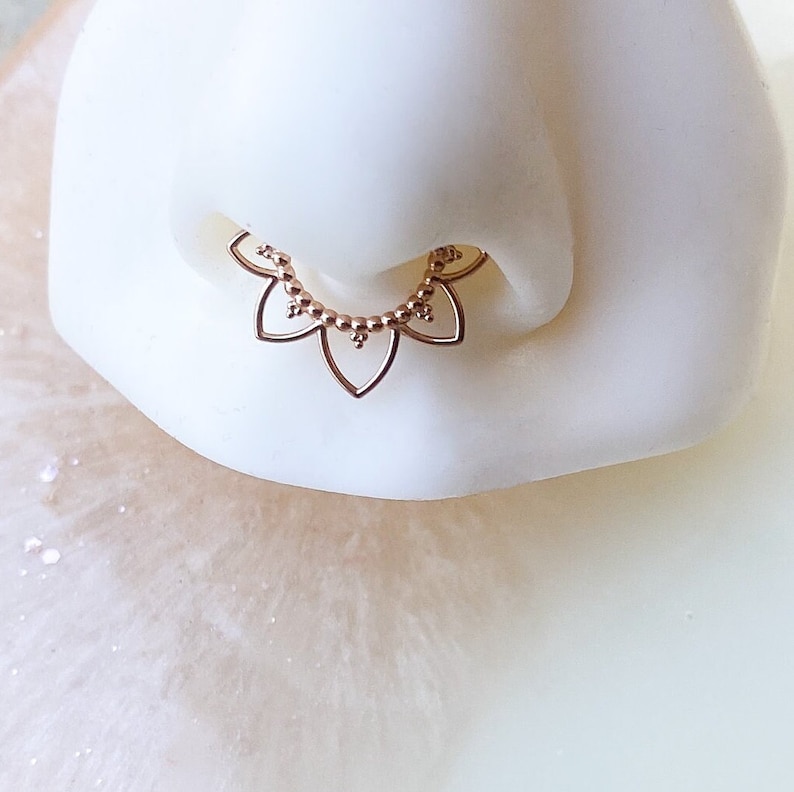 Op de afbeelding: Ros&eacute;gouden septumring met een bloemmotief. De sieraden bestaan uit een reeks bloemblad-vormige elementen en kleine, ronde kralen. De ring is ontworpen om in de neus te worden gedragen.
