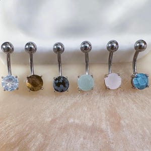 May include: Six belly button rings with different colored stones. The stones are clear, brown, gray, green, pink, and blue.