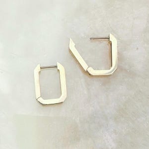 May include: Two gold-colored, geometric hoop earrings. Each earring has a rectangular shape with a post closure. The earrings are displayed on a light-colored, speckled surface. The earrings are a modern jewelry design.