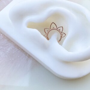Op de afbeelding: Ros&eacute;gouden septumring met een bloemmotief. De ring heeft een reeks kleine, met kralen bezette accenten en bloembladvormige uitsparingen. De sieraden worden gepresenteerd op een wit, getextureerd oppervlak.