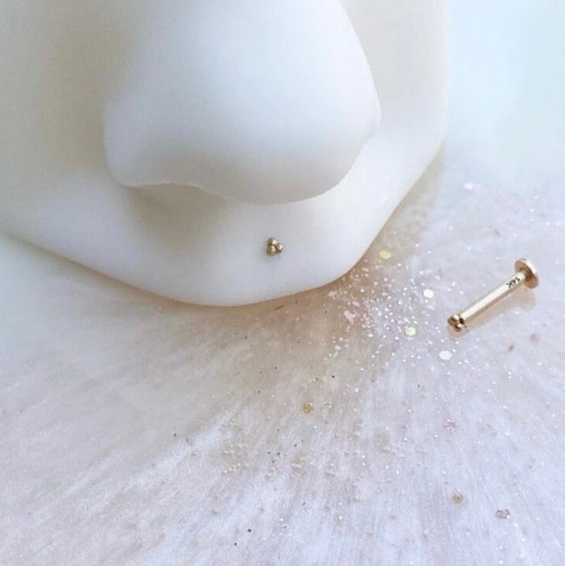 May include: A gold nose stud with three small balls on the end. The stud is lying on a white surface with glitter.
