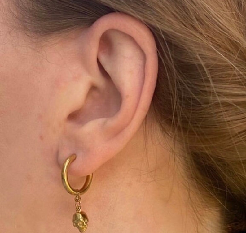 May include: Gold hoop earrings with a small gold skull charm dangling from the bottom.
