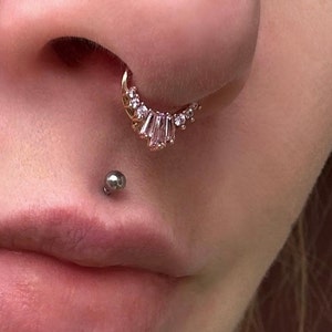 Pode incluir: Um close-up do rosto de uma pessoa com um piercing no nariz e um piercing no l&aacute;bio. O piercing no nariz &eacute; um aro de ouro com uma fileira de pequenas pedras transparentes. O piercing no l&aacute;bio &eacute; uma pequena bola de prata.
