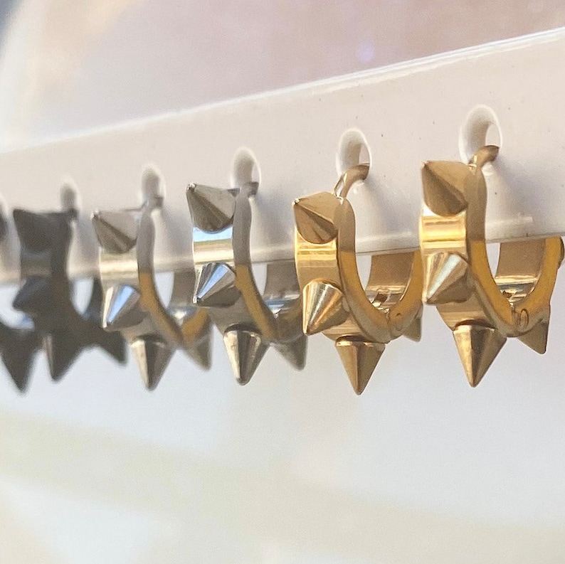 May include: A set of five hoop earrings with a row of spikes around the hoop. The earrings are available in black, silver, and gold.
