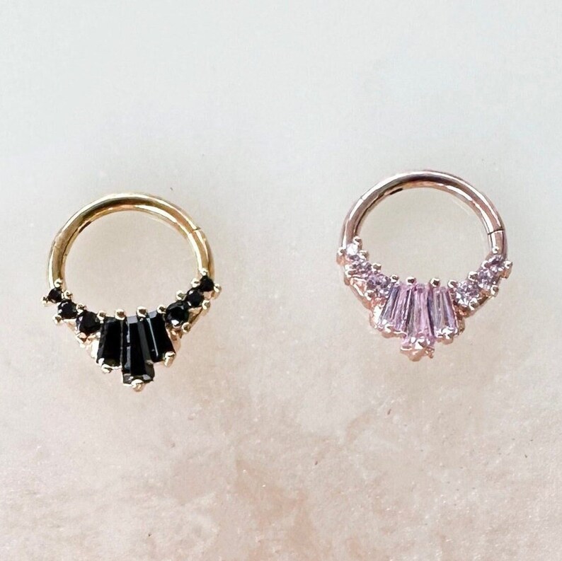 Pode incluir: Dois brincos de argola de ouro com uma fileira de pedras pequenas e uma pedra maior no centro. O brinco esquerdo tem pedras pretas e o brinco direito tem pedras cor-de-rosa.