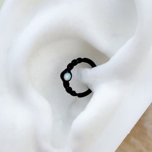 May include: Black hoop earring with a small opal gemstone. The earring is shown in a white background.