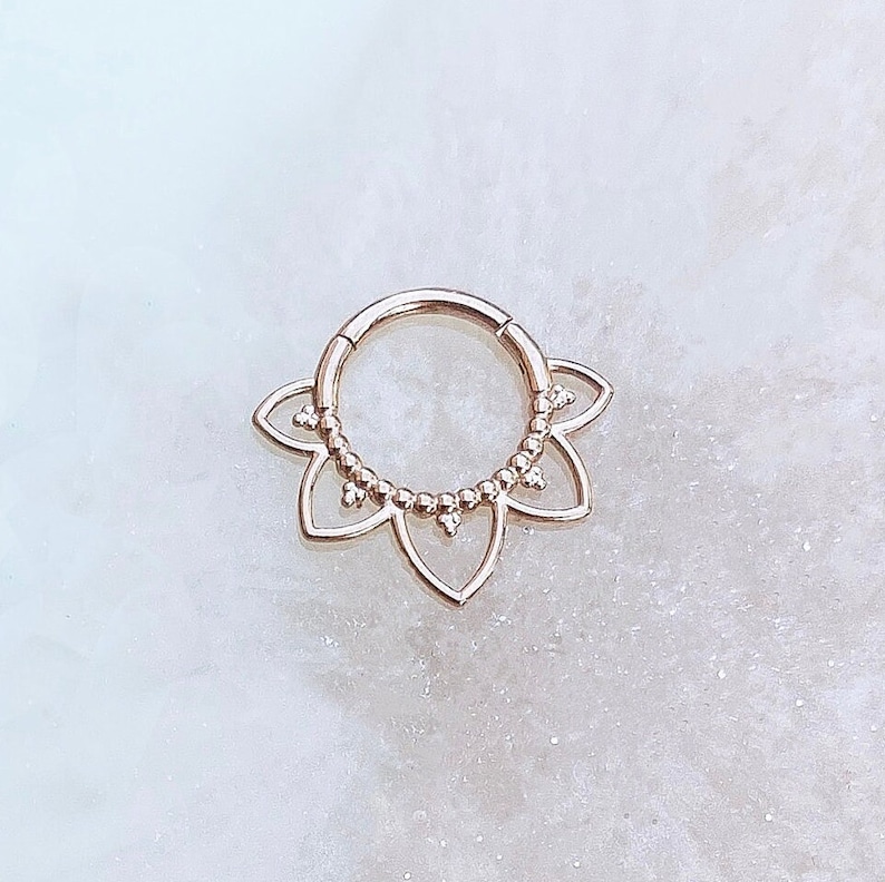 Op de afbeelding: Rosegouden septum clicker ring met een bloemmotief. De ring heeft een reeks kleine kralen en een bloemvorm. De sieraden zijn ontworpen voor body piercing.