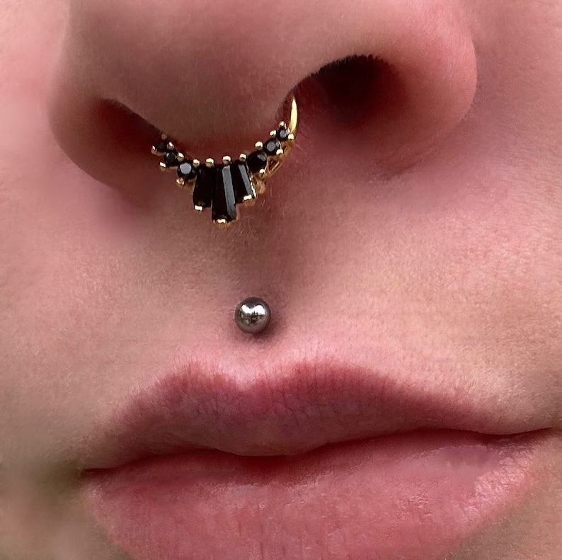Pode incluir: Um close-up do rosto de uma pessoa com dois piercings. O piercing no nariz &eacute; um aro de ouro com pedras pretas, e o piercing no l&aacute;bio &eacute; uma pequena bola de prata.