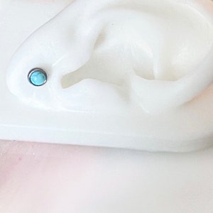 May include: A white plastic ear model with a turquoise gemstone stud earring in the helix piercing.