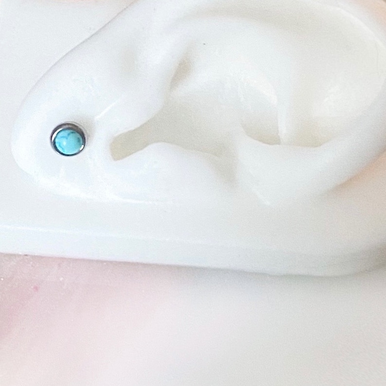 May include: A white plastic ear model with a turquoise gemstone stud earring in the helix piercing.