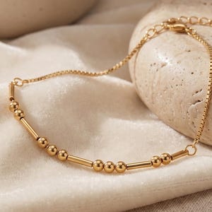 May include: A gold-toned chain bracelet featuring a series of small spherical beads and short cylindrical bars arranged in a repeating pattern. The bracelet includes a delicate box chain design and an adjustable clasp mechanism at the end.