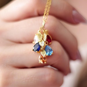 May include: A gold necklace with a charm featuring multiple teardrop-shaped gemstones in blue, red, and clear. The charm is adorned with small gold leaves.