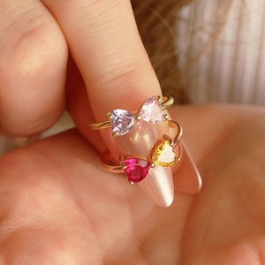 May include: Four gold rings, each with a heart-shaped gemstone in shades of pink, purple, and yellow. The rings are displayed on a finger and in the palm of a hand, showcasing the jewelry's design and color variations.