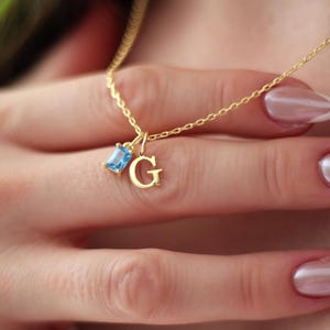 May include: A gold-colored necklace with a small, light blue gemstone and the letter "G" pendant. The necklace is delicate and appears to be made of a fine chain. The gemstone is rectangular. The necklace is displayed on a person.
