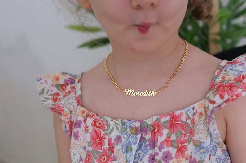 Pu&ograve; includere: Una giovane persona che indossa un vestito floreale con una collana a catena dorata che dice "Meredith".