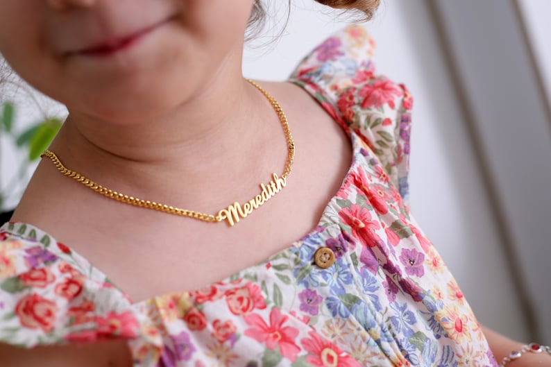Pu&ograve; includere: Una collana a catena d'oro con il nome "Meredith" scritto in corsivo. La collana &egrave; indossata da un bambino che indossa un vestito floreale.