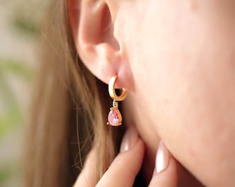May include: A gold hoop earring with a pink teardrop-shaped gemstone. The earring is worn on the ear of a person. The gemstone is a light pink color and is set in a gold bezel.