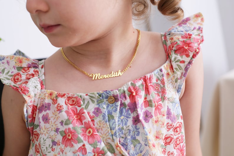 Pu&ograve; includere: Una giovane persona che indossa un vestito bianco con un motivo floreale e una collana d'oro con il nome "Meredith" su di essa.