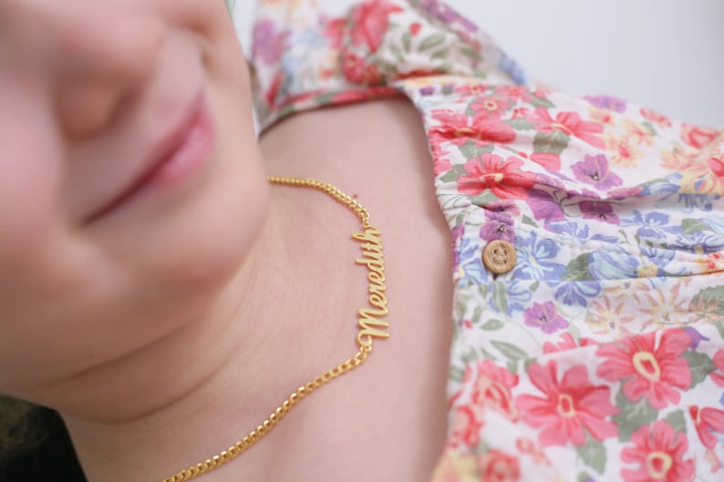 Pu&ograve; includere: Una collana a catena dorata con il nome "Meredith" scritto in corsivo. La collana &egrave; indossata da una persona con una camicia a fantasia floreale.