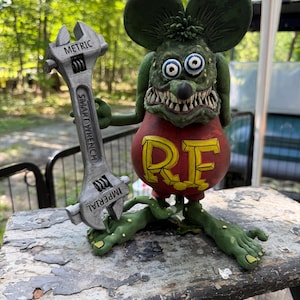 May include: A green, cartoon-style mouse figurine with large ears, bulging eyes, and sharp teeth, wearing a red shirt with the yellow letters RF. The mouse holds a silver-coloured wrench labelled METRIC, SMARTWRENCH, and IMPERIAL.