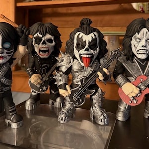 以下が含まれることがあります： ロックバンドKISSの4体のコレクターズフィギュア。各フィギュアは、詳細な顔のペイントとコスチュームをしています。1体はドラムスティックを持ち、他はギターとベースを持っています。フィギュアは黒、シルバー、赤です。