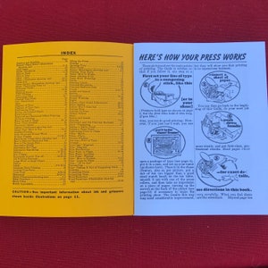 May include: A yellow and white guidebook with the title "Here's How Your Press Works" and illustrations showing how to use a printing press. The text includes instructions on how to insert a sheet of paper, set the type, and push down the handle.