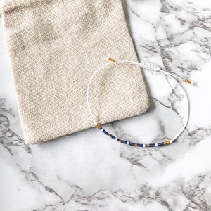 Könnte beinhalten: Ein weißes Kordelarmband mit blauen und goldenen Perlen, neben einem beigen Kordelzugbeutel. Das Armband hat einen verstellbaren Knotenverschluss. Der Hintergrund ist ein weißes und graues Marmormuster.