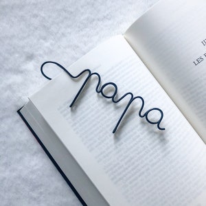 Peut inclure: Un marque-page en métal bleu foncé en forme du mot "papa" est inséré dans un livre ouvert. Les pages du livre sont blanches et le texte sur les pages est visible. Le marque-page est de conception simple et élégante.