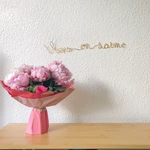 Peut inclure: Un bouquet de pivoines roses enveloppé dans du papier rose et rouge, posé sur une surface en bois. Au-dessus du bouquet, le texte en fil d'or dit "Viens en larme" contre un mur blanc texturé.