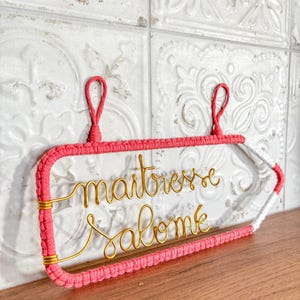 May include: A pink macrame sign with the words "maitresse salome" in gold wire. The sign has a clear center and a white and pink tail. It hangs from two pink loops.