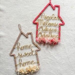 Peut inclure: Deux décorations murales en forme de maison. L'une est rose avec des noms en fil d'or : Nicolas, Mélanie, Adrien et Adaline. L'autre est beige avec les mots "home sweet home" en fil d'or. Les deux ont des fleurs séchées.