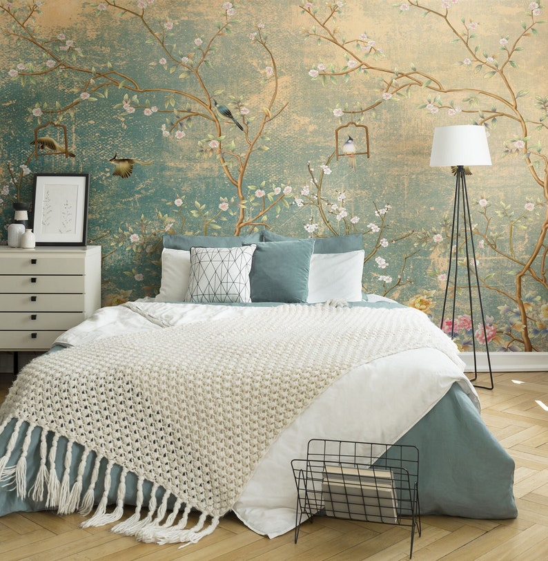 May include: A bedroom with a teal blue and beige floral wallpaper featuring birds and flowers. The bed has a white knitted throw blanket with fringe and a teal blue duvet. A black metal magazine rack is on the floor next to the bed. A white lamp with a black tripod stand is in the corner of the room.