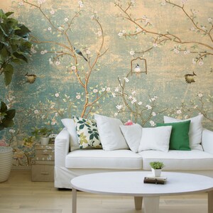 May include: A white sofa with green and floral patterned throw pillows sits in front of a wall with a vintage floral wallpaper featuring birds and a birdcage. A small potted plant sits on a white coffee table in front of the sofa.