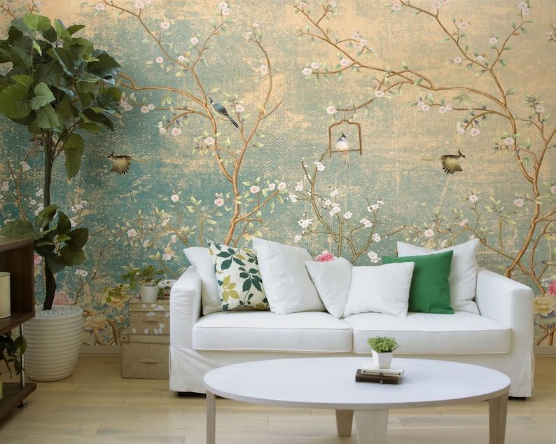May include: A white sofa with green and floral patterned throw pillows sits in front of a wall with a vintage floral wallpaper featuring birds and a birdcage. A small potted plant sits on a white coffee table in front of the sofa.