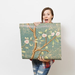 May include: A woman holds up a roll of wallpaper with a floral pattern. The wallpaper is blue with gold branches and pink flowers. The woman is wearing a red and black plaid shirt and blue jeans.