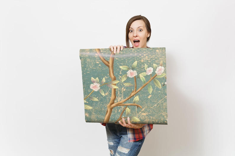 May include: A woman holds up a roll of wallpaper with a floral pattern. The wallpaper is blue with gold branches and pink flowers. The woman is wearing a red and black plaid shirt and blue jeans.