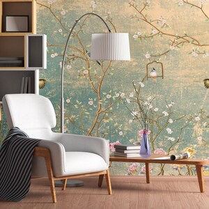May include: A white armchair with a black and white striped throw blanket sits in a room with a floral wallpaper featuring birds and branches. A floor lamp with a white shade and a vase with white flowers are also in the room.
