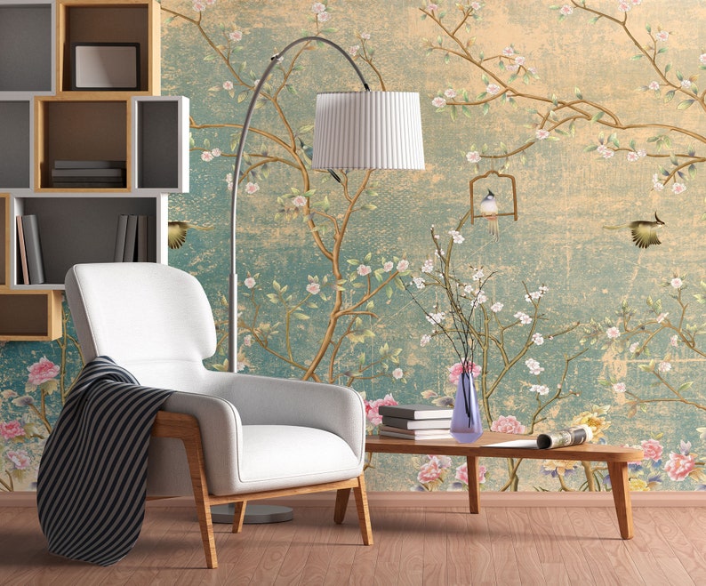May include: A white armchair with a black and white striped throw blanket sits in a room with a floral wallpaper featuring birds and branches. A floor lamp with a white shade and a vase with white flowers are also in the room.