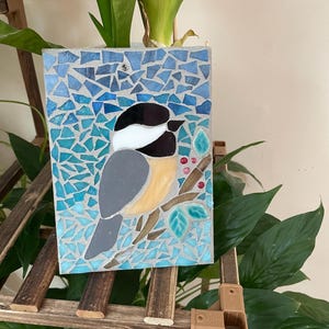 May include: A mosaic art piece featuring a bird perched on a branch with leaves and berries. The bird has a black and white head, gray body, and yellow chest. The background is a mosaic of blue tiles. The artwork is displayed on a wooden easel.