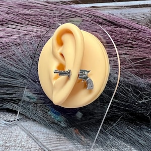 May include: A close-up of a fake ear with two silver gun-shaped earrings. The earrings are attached to the ear with a stud and a bar.