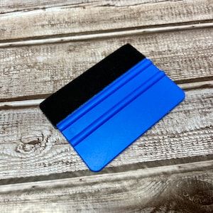 Puede incluir: Una escobilla de goma de plástico azul y negro con un borde de fieltro. La escobilla tiene tres líneas elevadas en el lado azul.