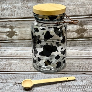 Puede incluir: Un tarro de vidrio transparente con un diseño de vaca en blanco y negro. El tarro tiene una tapa de madera y un asa de cuerda marrón. Se incluye una cuchara de madera.