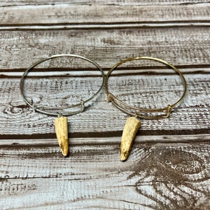 May include: Two adjustable bangle bracelets. One is silver-colored, the other is gold-colored. Each bracelet has a pendant made of a light brown antler. The bracelets are displayed on a light brown wooden surface.